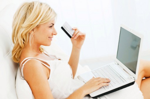 Cheerful blonde woman sitting in her bed and shopping online with credit card and laptop computer.